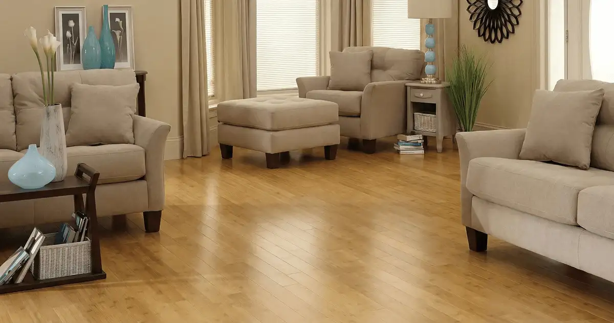 A living room with professionally installed bamboo flooring, beige sofas, a matching ottoman, a side table with a blue lamp, vases, framed pictures, and large windows with blinds and curtains in Buffalo, NY